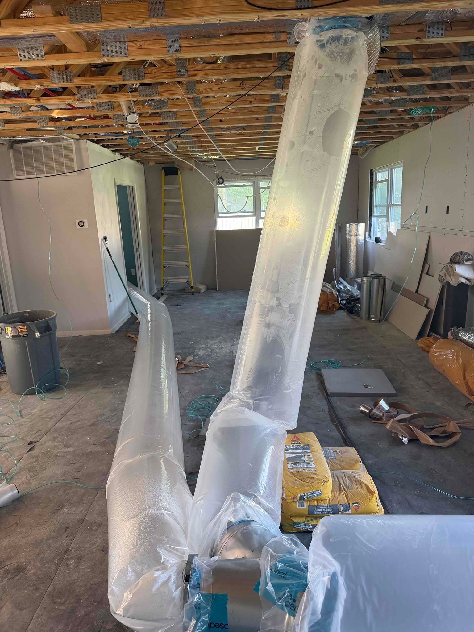 Renovation room with exposed ceiling and walls, plastic-wrapped ducting, ladders, and scattered tools.