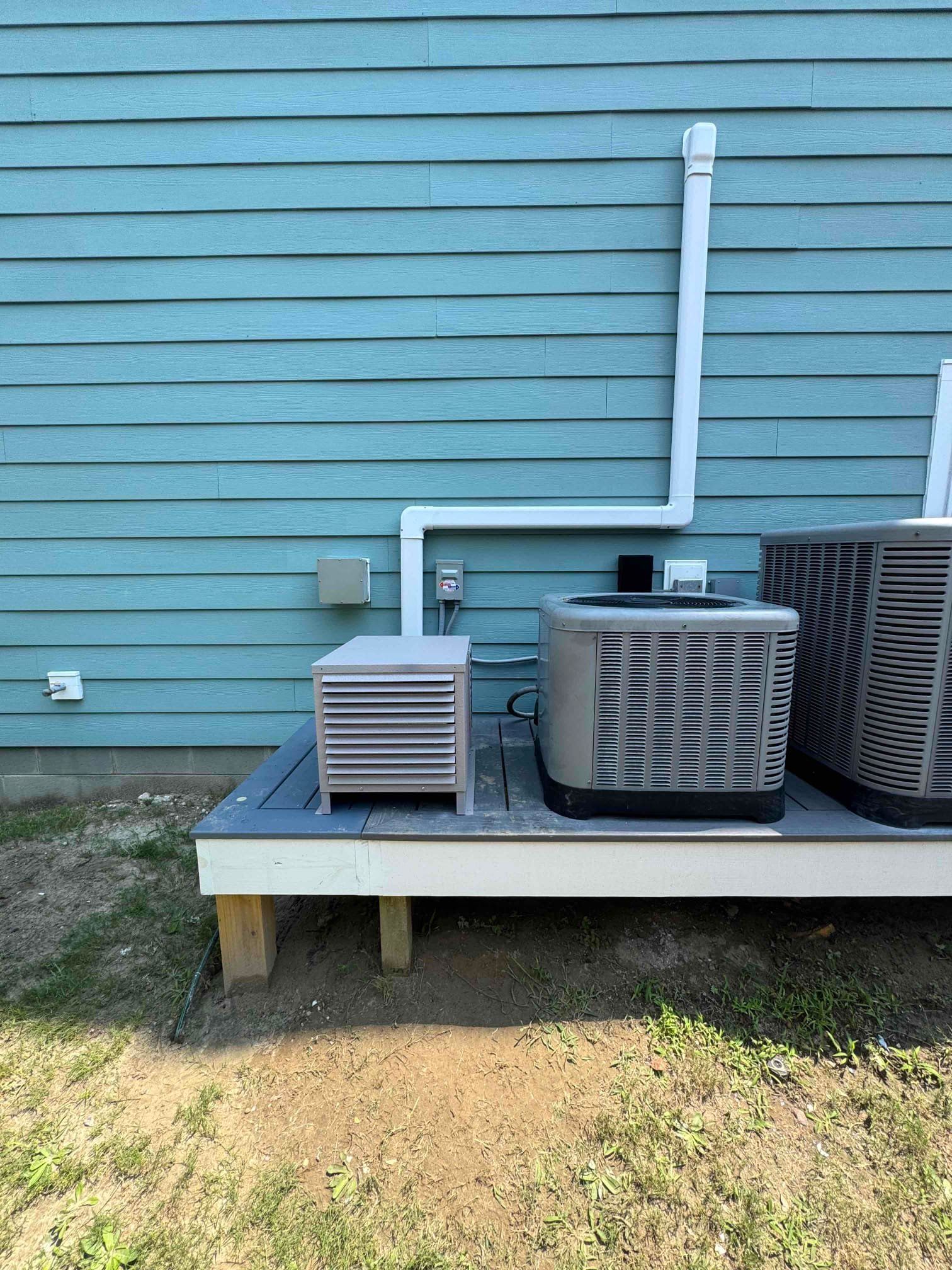 Outdoor HVAC units on a raised platform against a blue house wall with white piping and conduit.