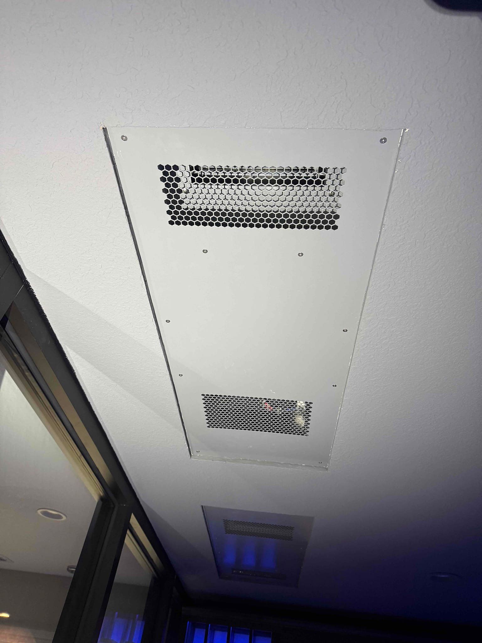 White ceiling vent panel with two black grilles in an indoor hallway near a window.