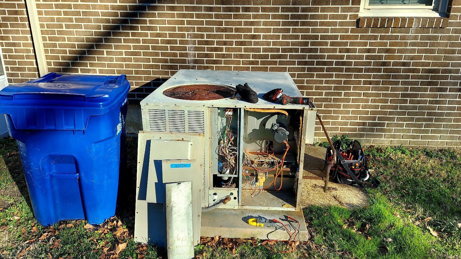 Blue trash bin beside a damaged outdoor HVAC unit against a brick wall.