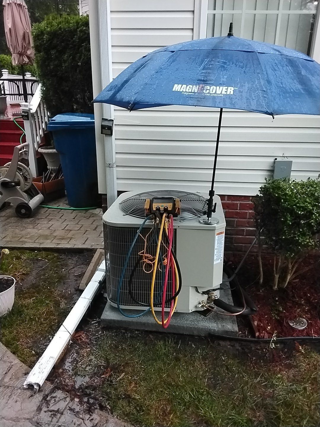 Blue umbrella covering an outdoor air conditioner unit beside a house and garden beds.