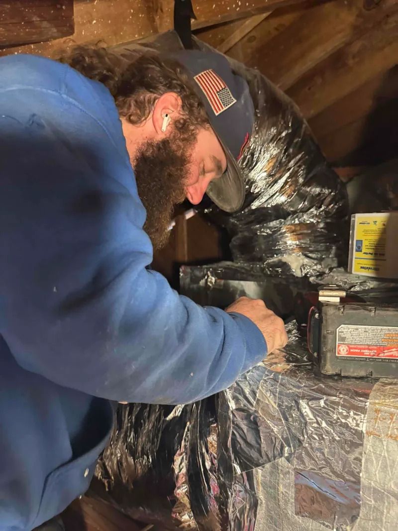 Worker in blue hoodie and cap inspects a wrapped pipe connection in a utility space.