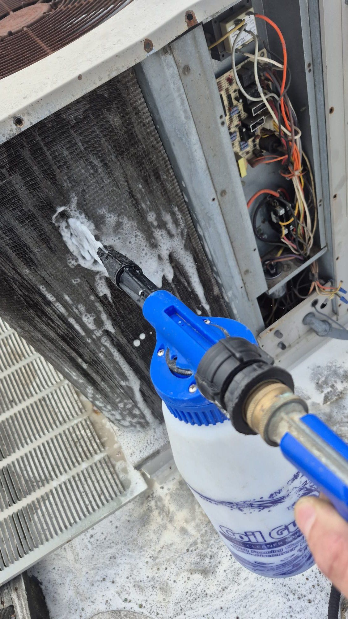 Blue spray nozzle cleaning a dirty air conditioner coil with foamy cleaner and a white bottle below.