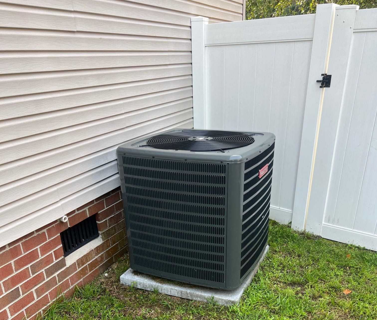 Outdoor air conditioner unit beside a house and white fence on a grassy yard.