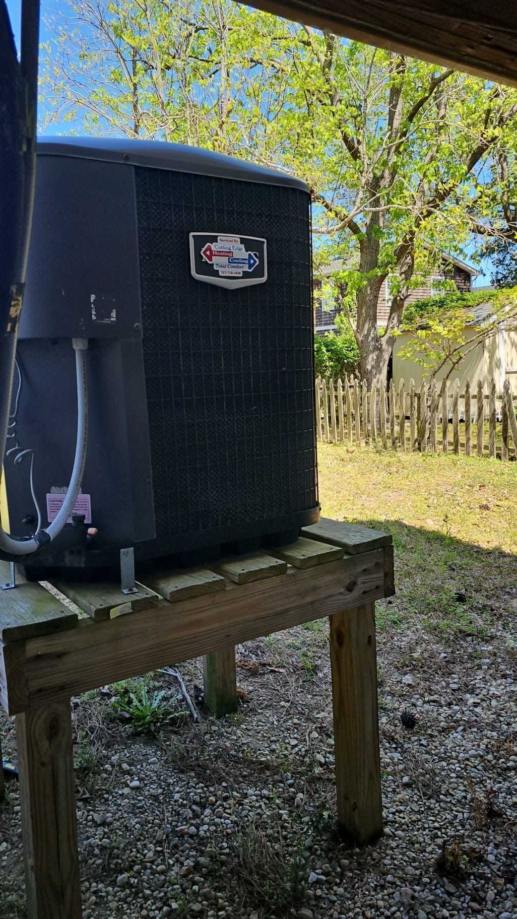 Black outdoor HVAC unit on a wooden platform in a yard, with trees and a fence in the background