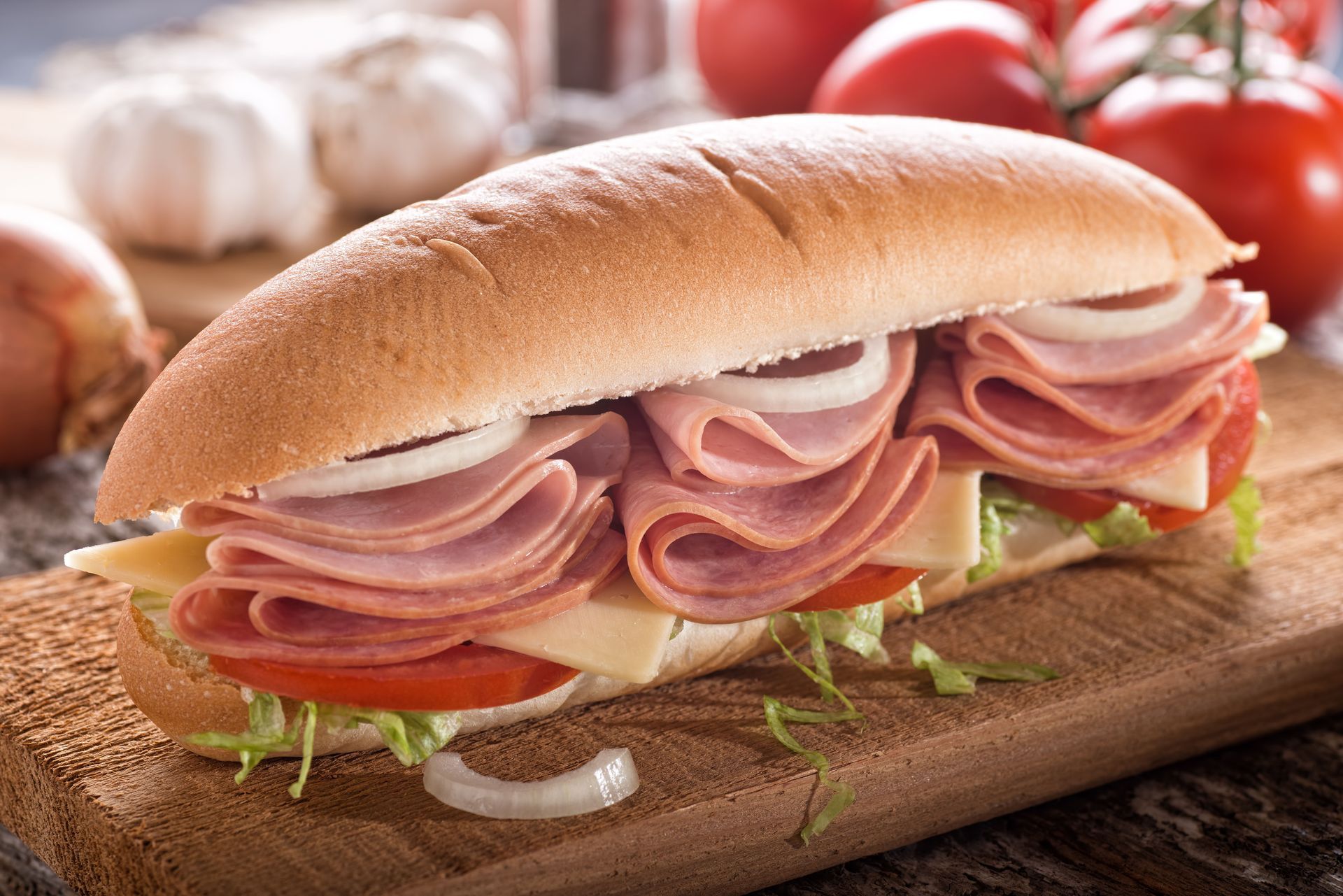 A sub sandwich with ham , cheese , tomatoes and onions on a wooden cutting board.