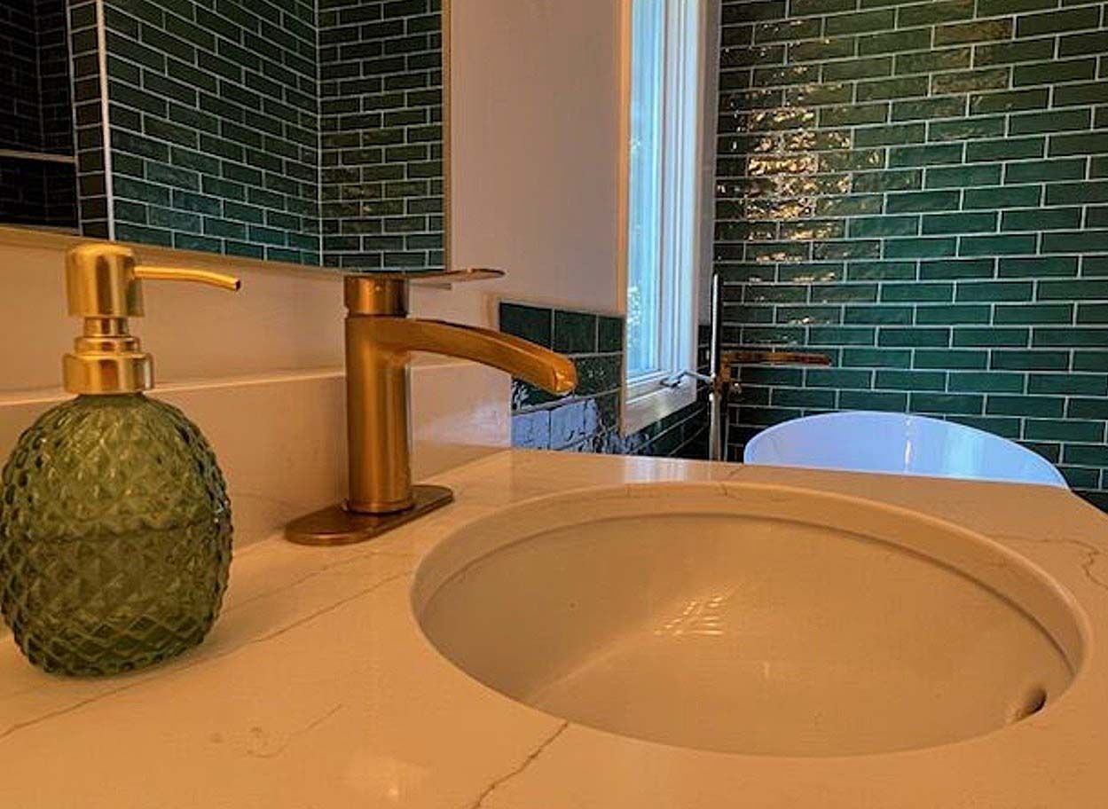 Bathroom sink with brass faucet, green soap dispenser, and green tile backsplash outside a window.