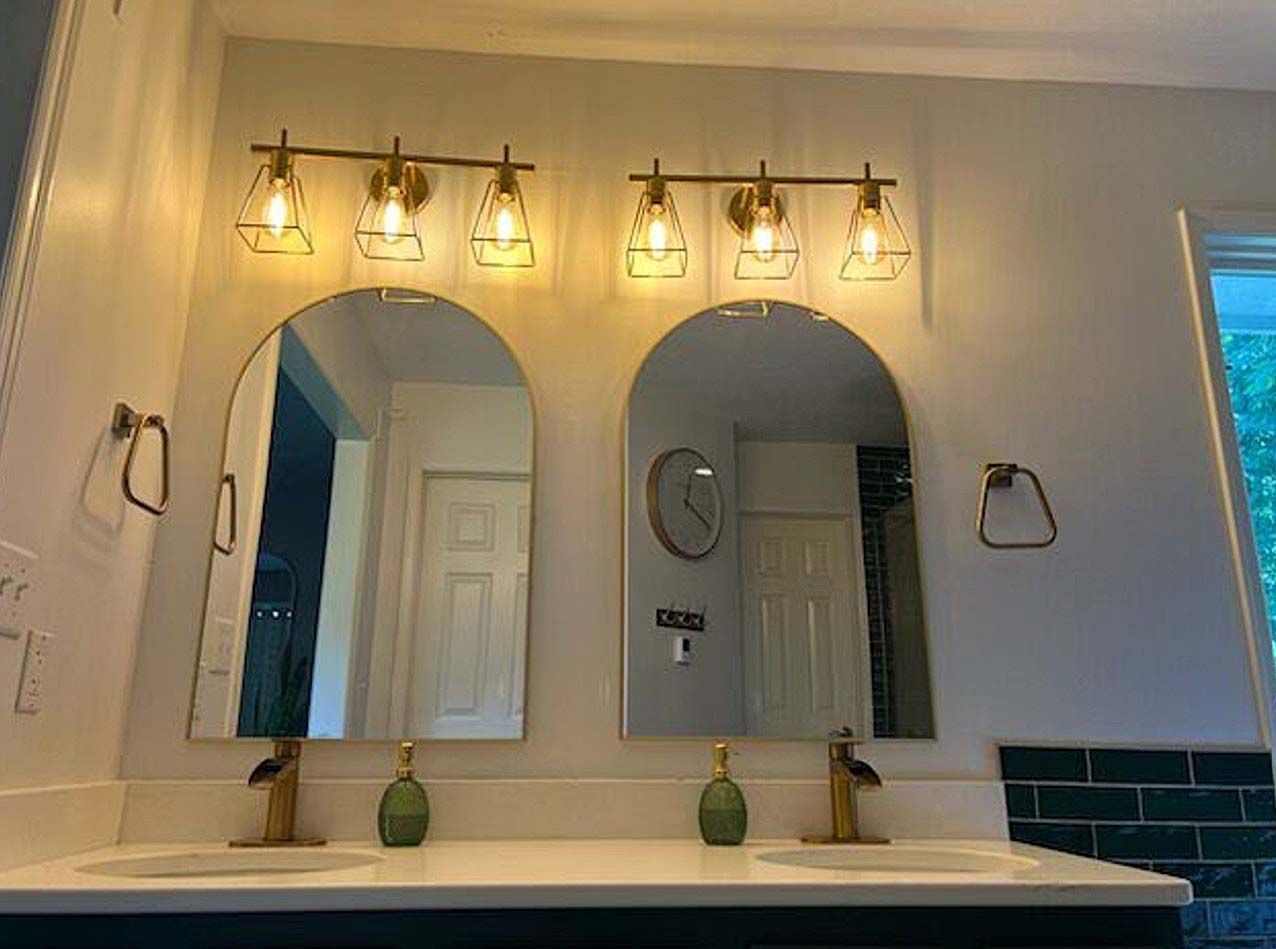 Two arched bathroom mirrors above a white double sink with brass faucets and warm vanity lights.