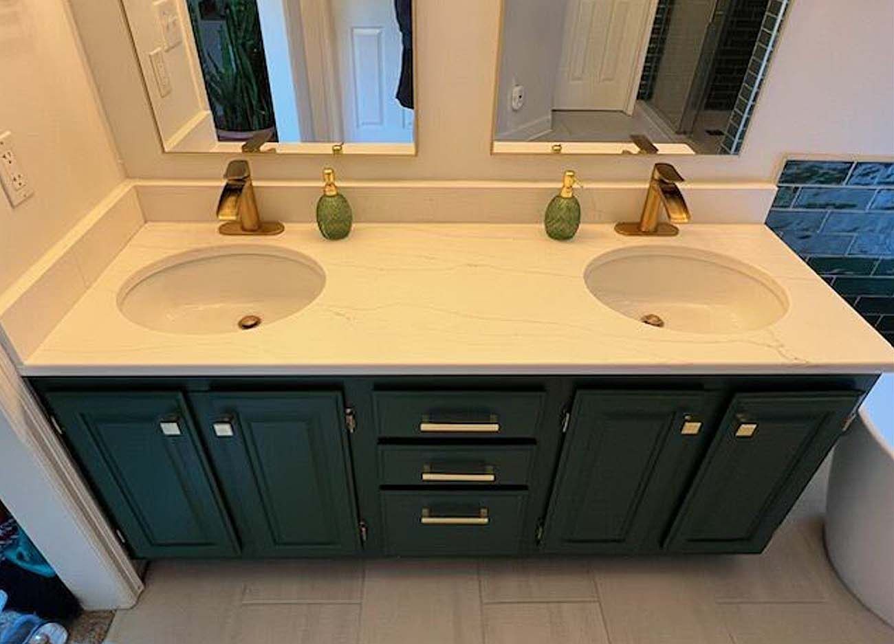 Double vanity bathroom sink with dark teal cabinets and two brass faucets under a mirror.