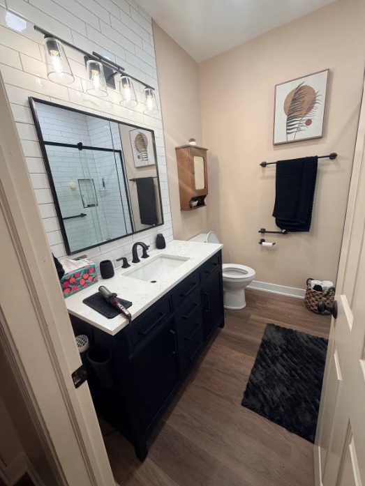 Small bathroom with black vanity, large mirror, white sink, toilet, towel rack, and bath mat