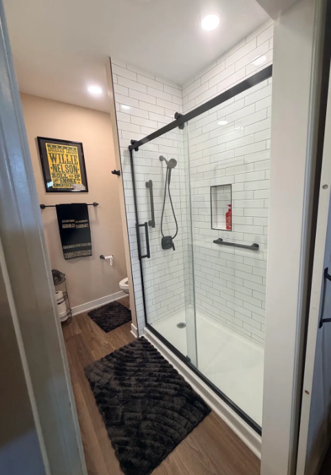 Narrow bathroom with a glass shower enclosure, white tile walls, wood floor, and dark rugs.
