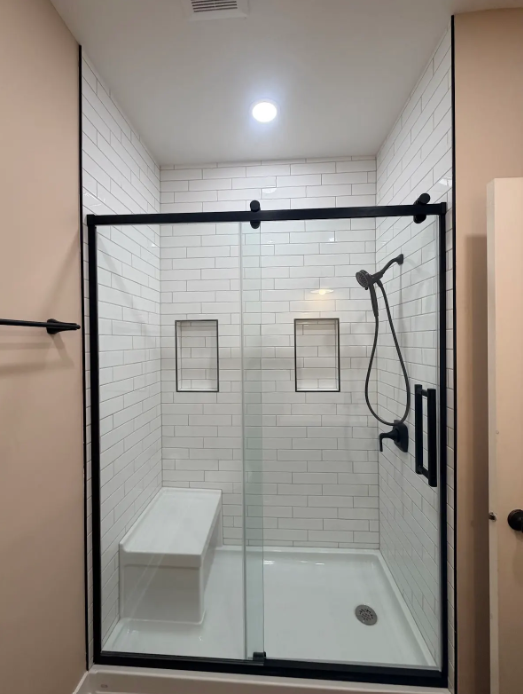 Modern walk-in shower with black-framed glass, white tile walls, bench, and handheld showerhead