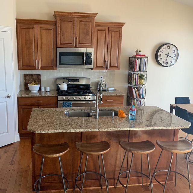 Kitchen with wooden cabinets, granite island, four stools, stainless stove, microwave, and wall clock.