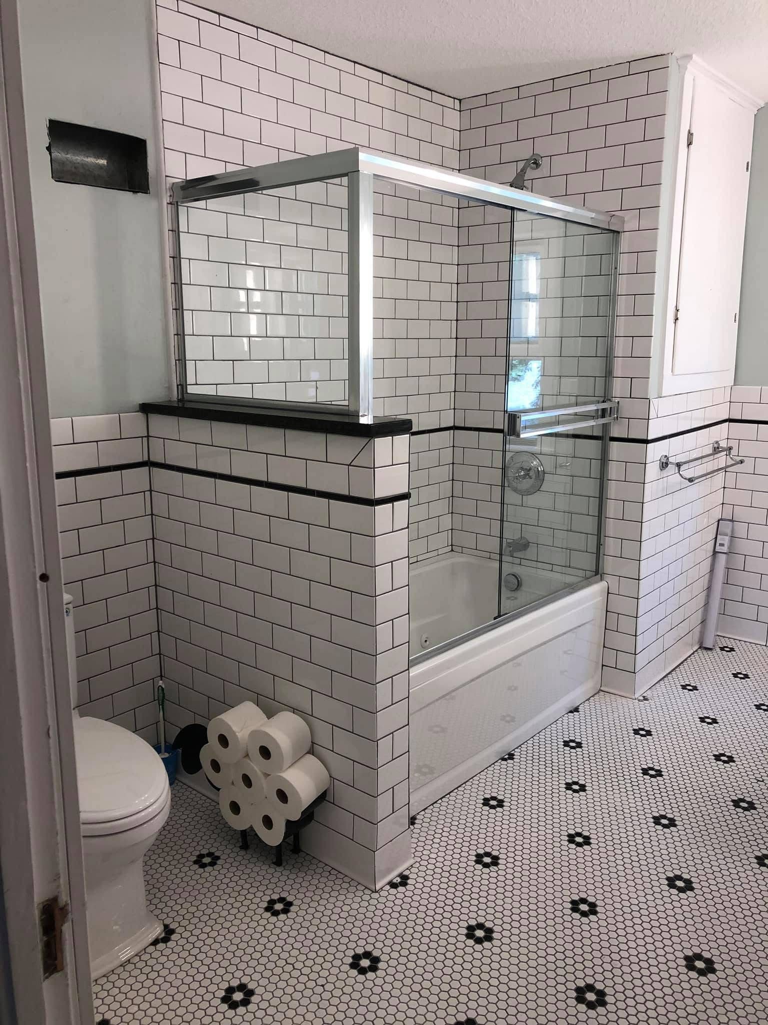 White-tiled bathroom with glass shower, toilet, and patterned black-and-white floor tiles.