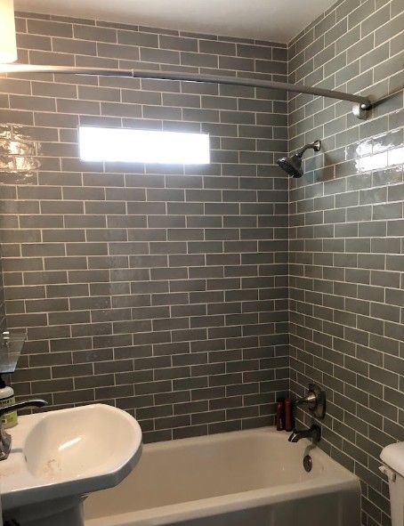 Bathroom with gray tile walls, bathtub, sink, window, and shower fixtures.
