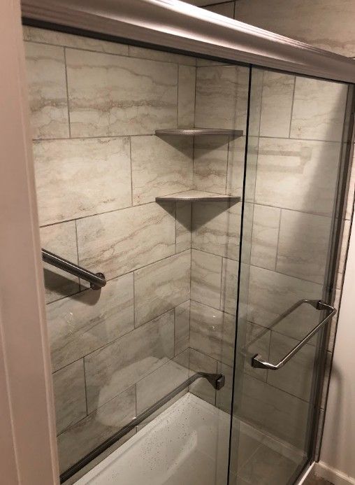 Glass shower with beige tile walls, built-in corner shelves, and a grab bar next to a bathtub.