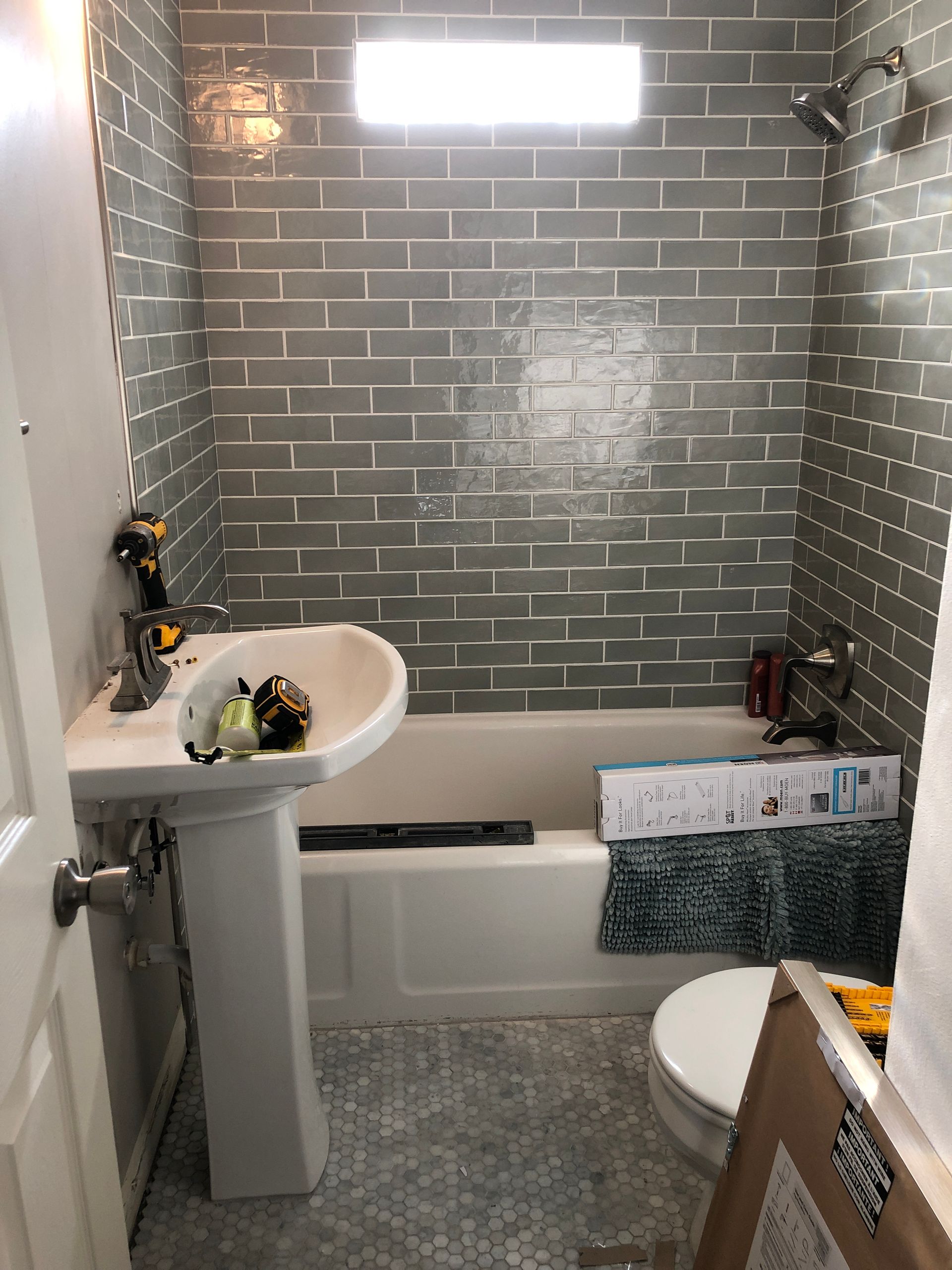 Small bathroom with a tub, pedestal sink, and toilet; gray tile walls and floor, bright window above tub.