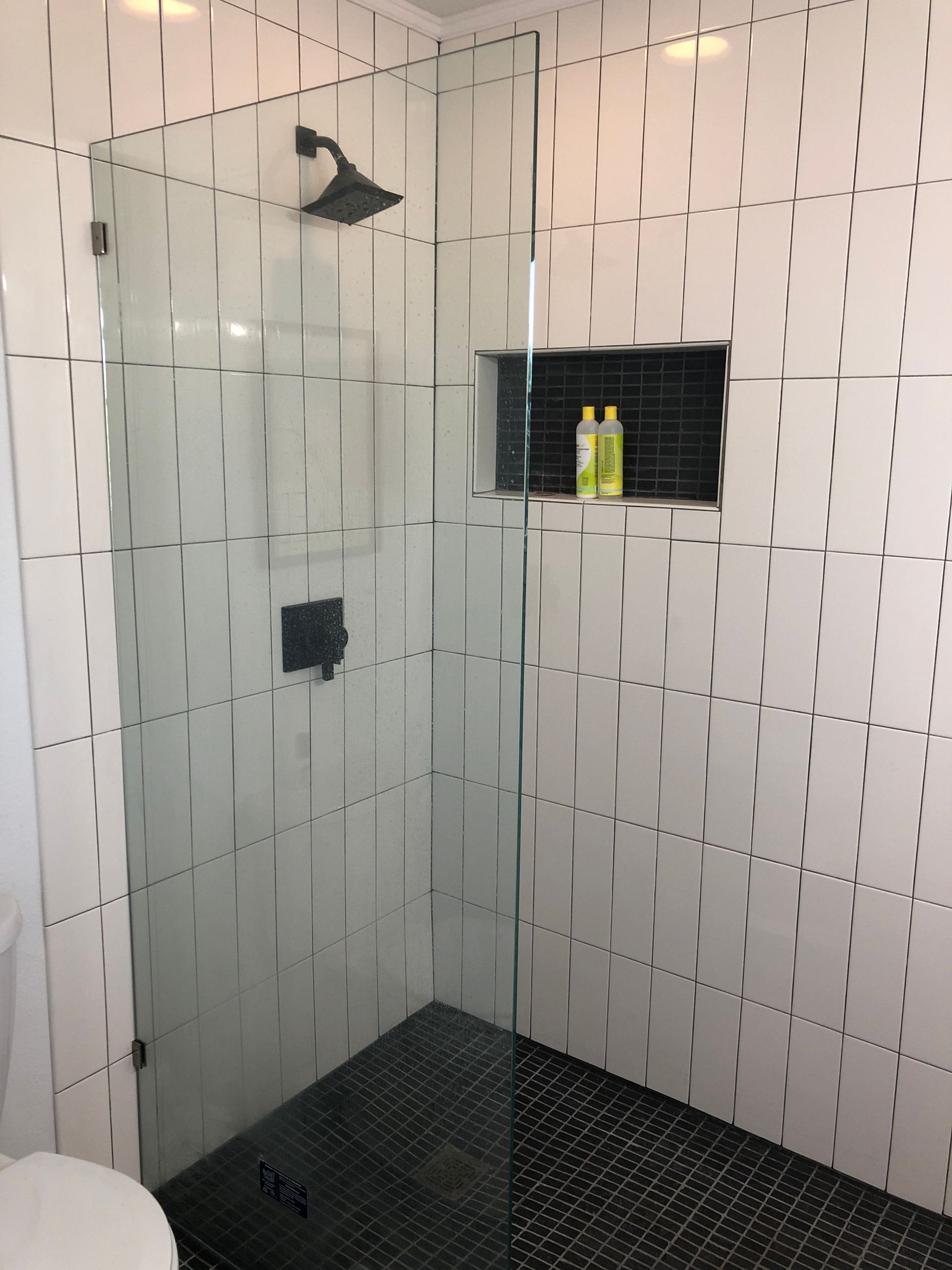 Tiled shower stall with a glass door, black shower floor, and recessed shelf holding bottles.