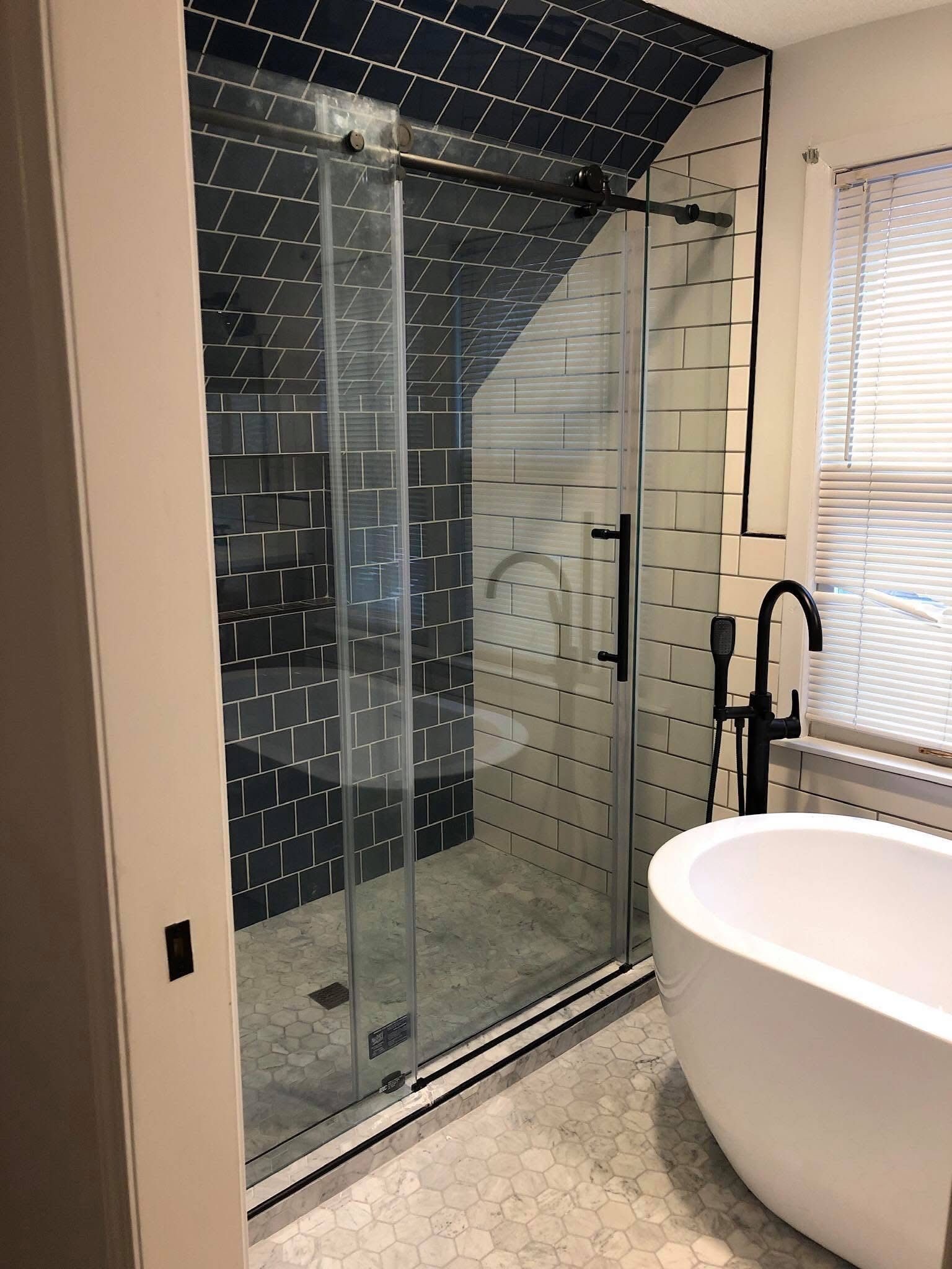 Modern bathroom with glass shower enclosure, black tile, and a white freestanding tub near a window.