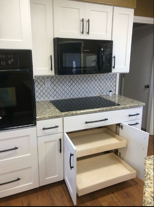 Kitchen with open lower cabinets, white cabinetry, black microwave, and stovetop under tiled backsplash.