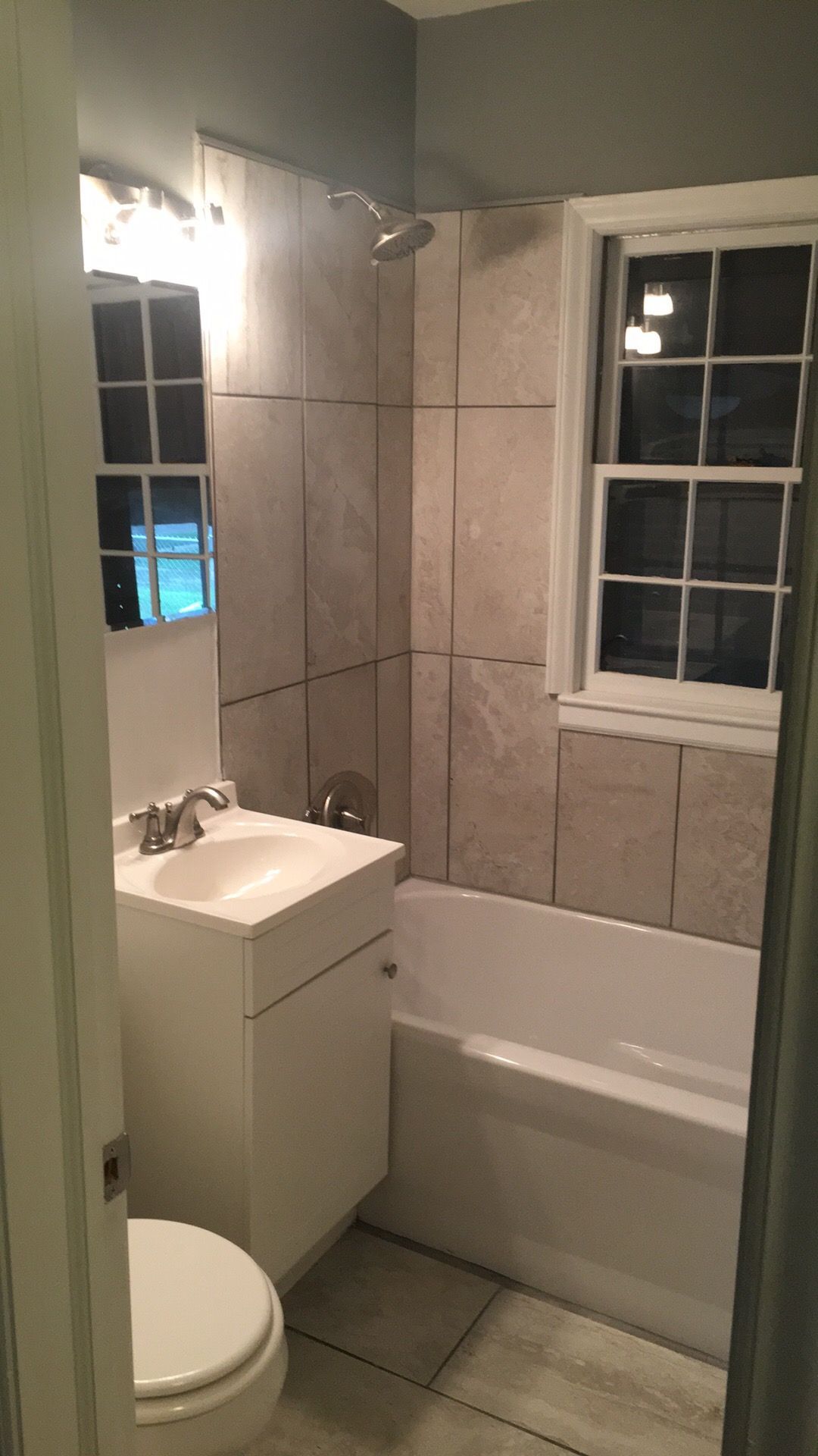 Small bathroom with white tub, sink vanity, toilet, and window beside a tiled shower wall.