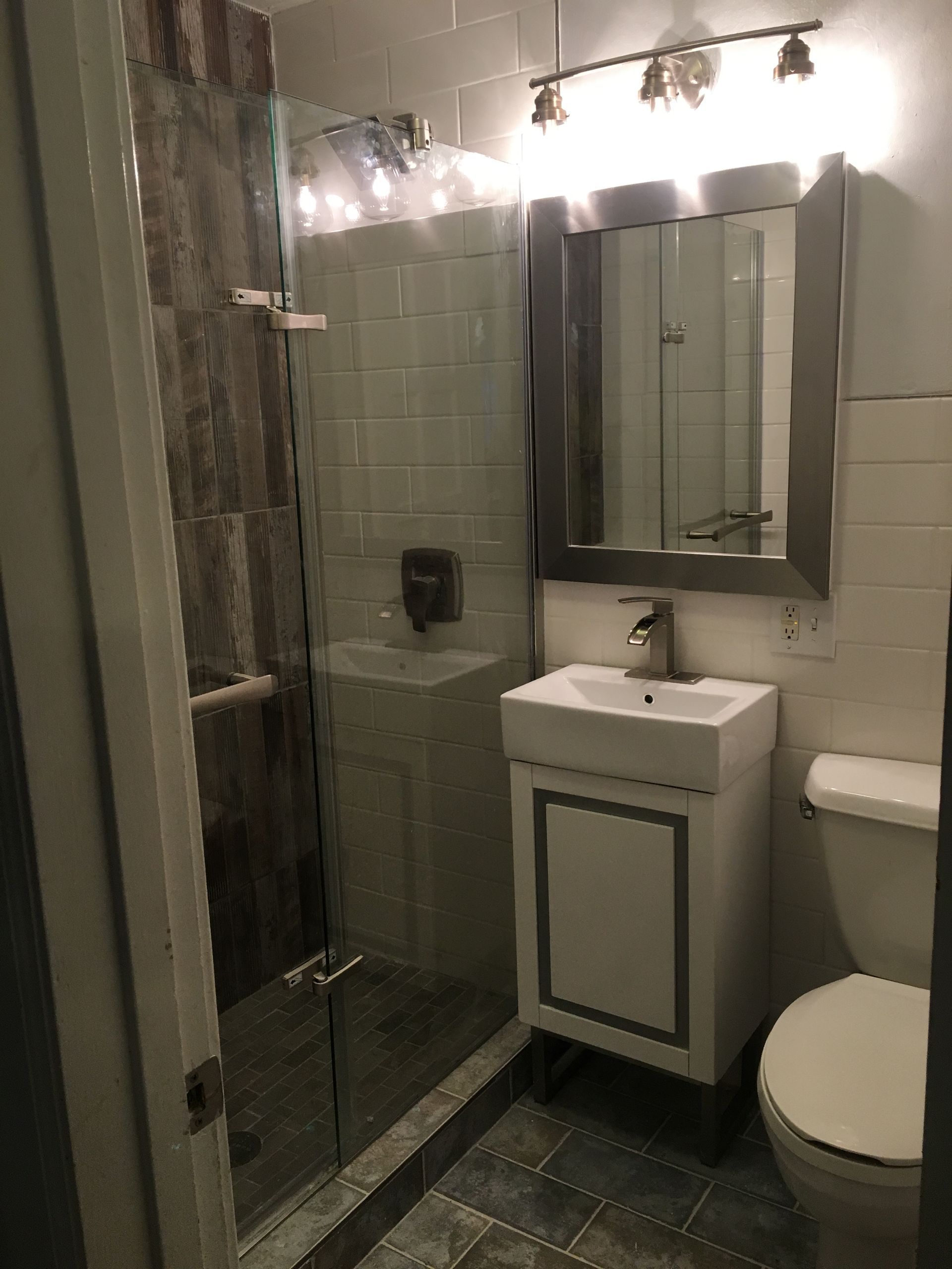 Small bathroom with glass shower, vanity sink, mirror, and toilet under bright lights.