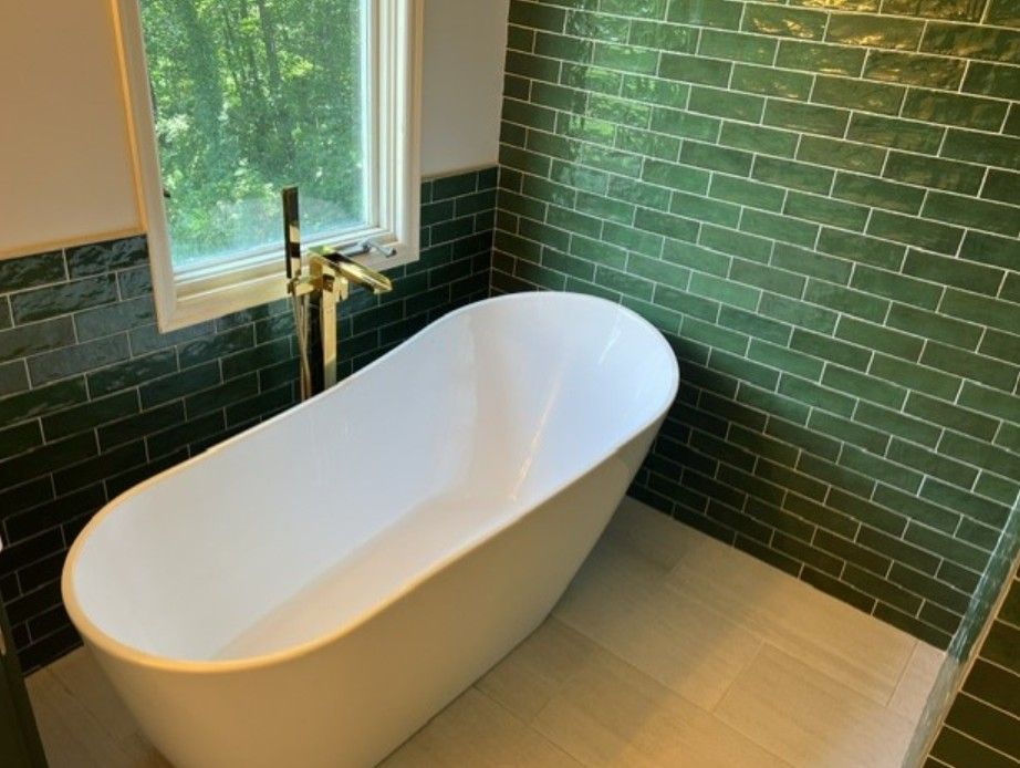 Modern bathroom with a freestanding white tub, brass faucet, green tiled walls, and a window.