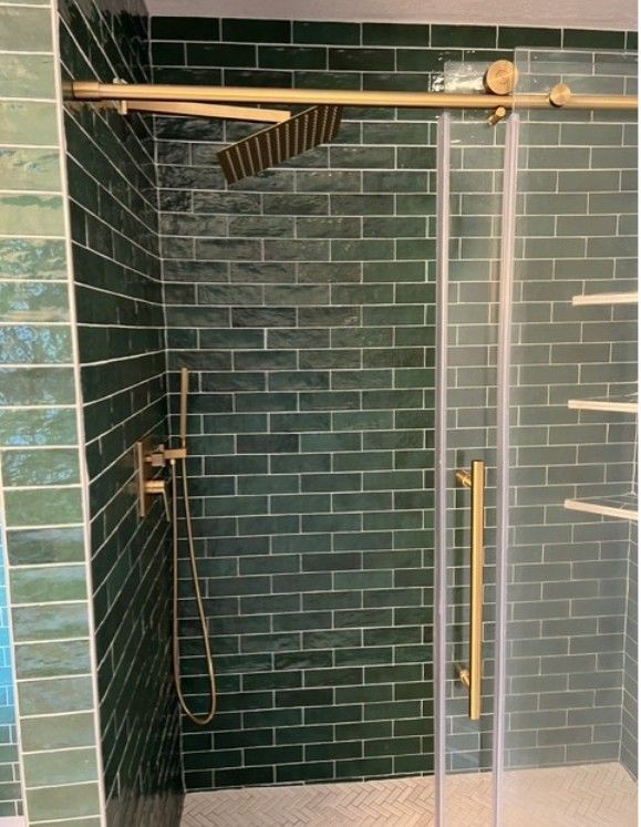 Walk-in shower with dark green tile walls, brass fixtures, glass door, and handheld showerhead.