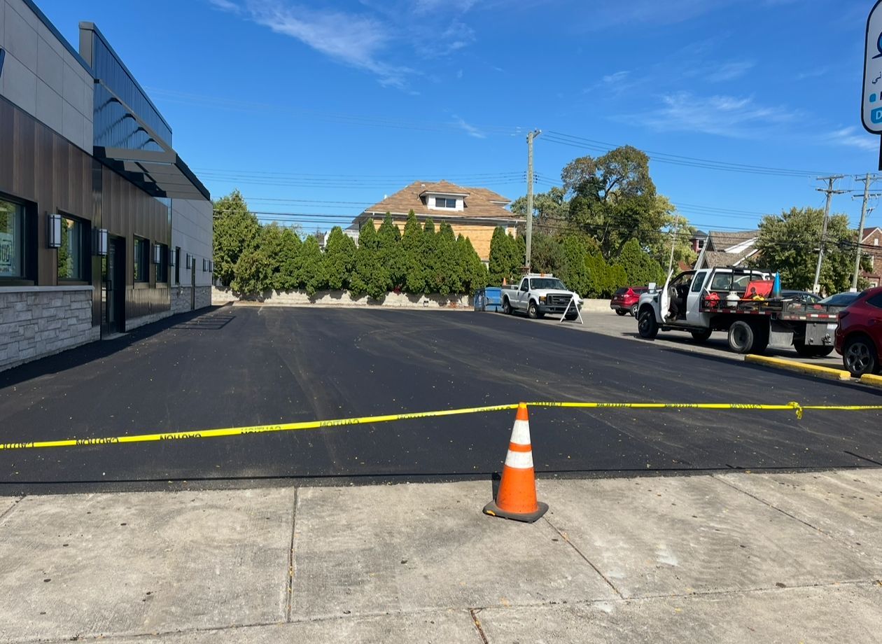 Freshly paved black asphalt parking lot blocked off by yellow caution tape and an orange traffic cone.