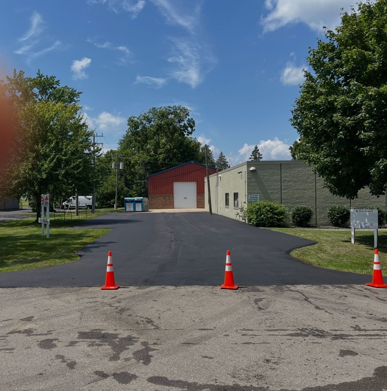A newly paved black asphalt driveway leads to a brick building, blocked by three orange traffic cones under a sunny sky.