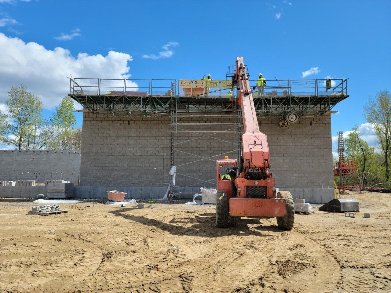 Construction workers on scaffolding with a telehandler lifting equipment near a brick wall under a blue sky.