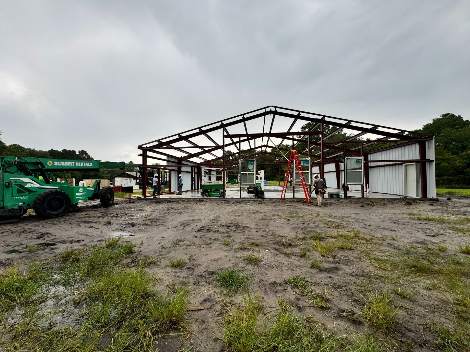 Construction of a metal building frame; muddy ground; green machinery and workers present.