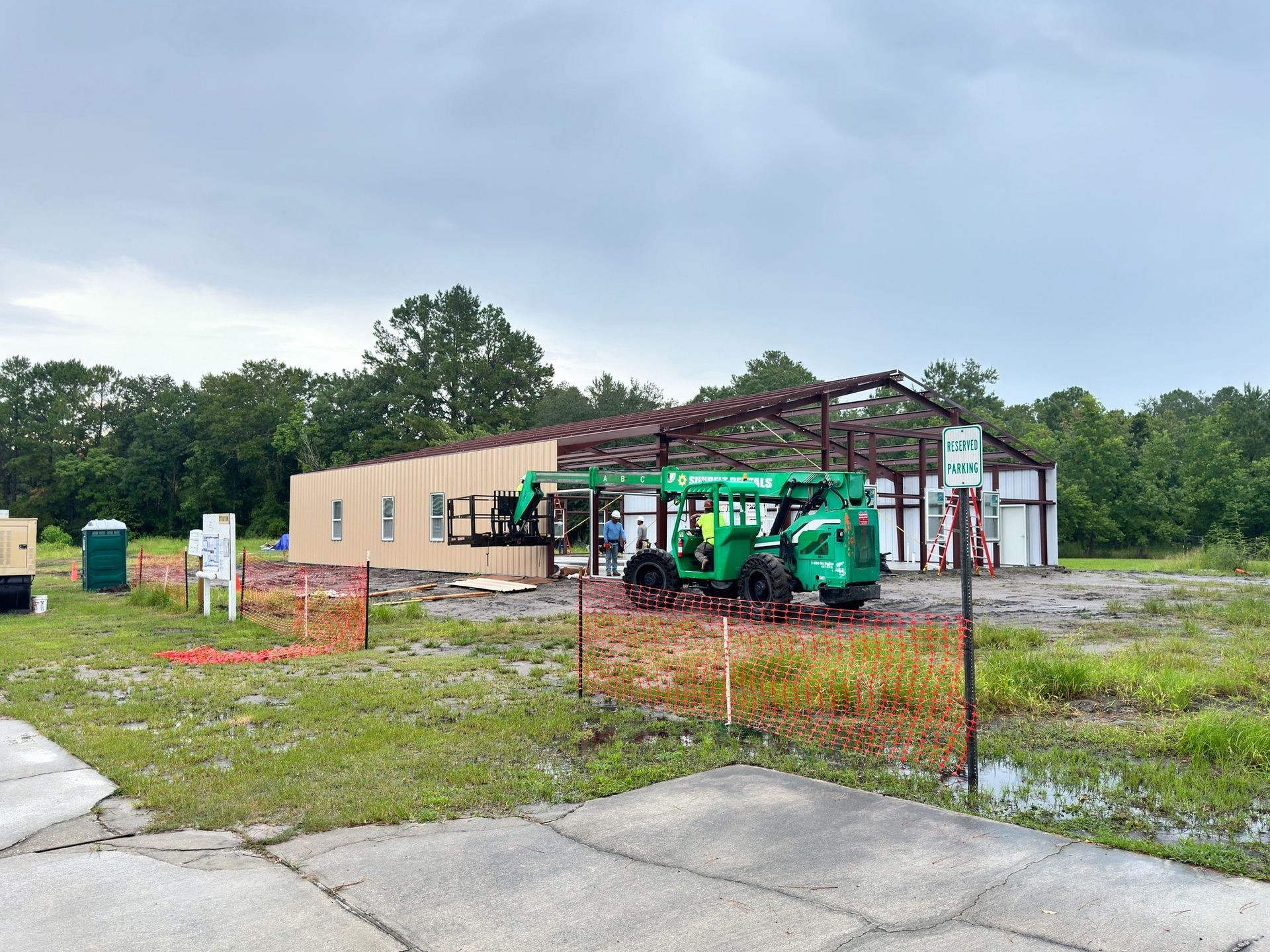 Construction of a tan and brown building with a green lift in front. Trees and overcast sky in the background.