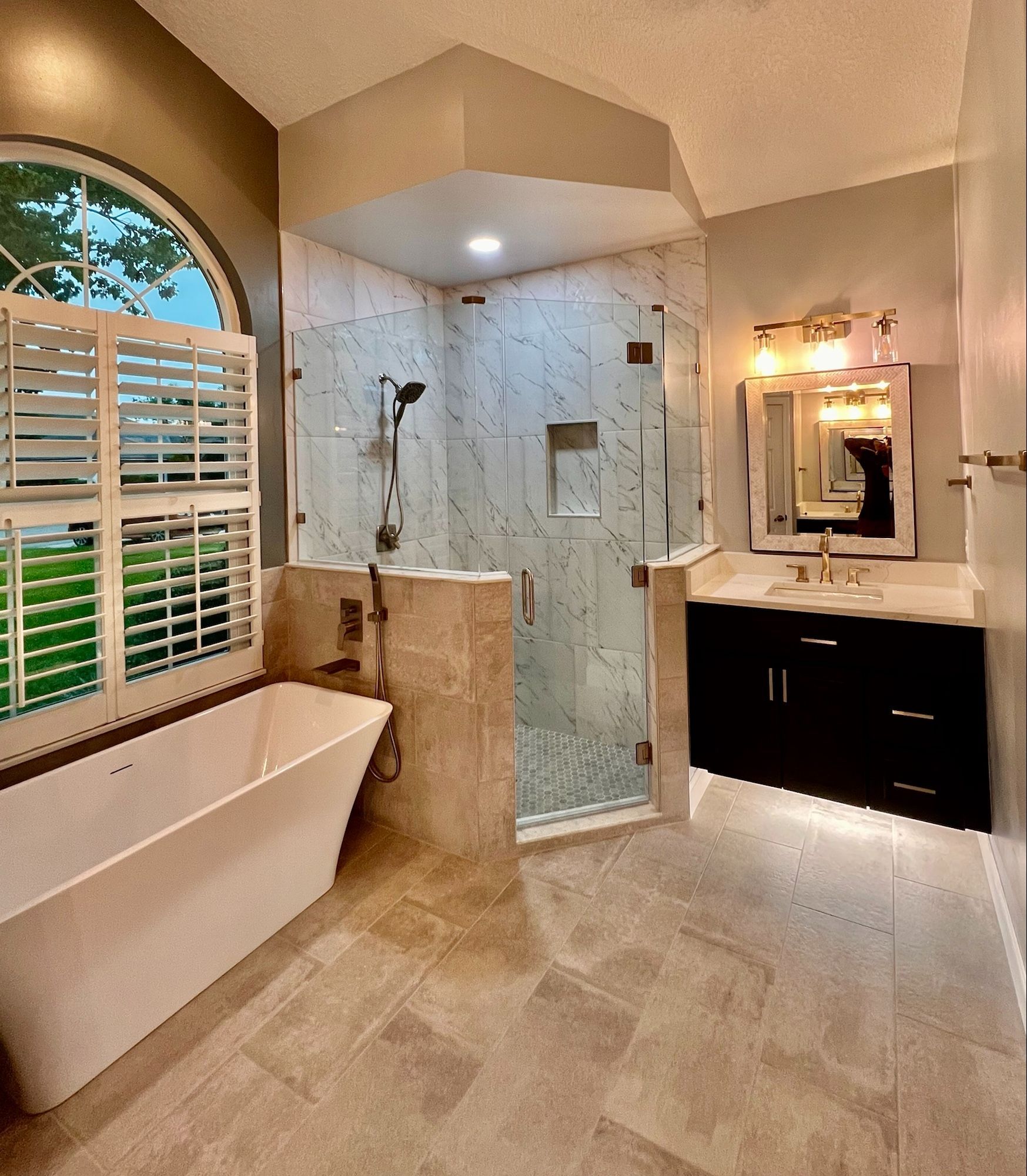 Spacious bathroom with glass shower, freestanding tub, dark vanity, and window with shutters.