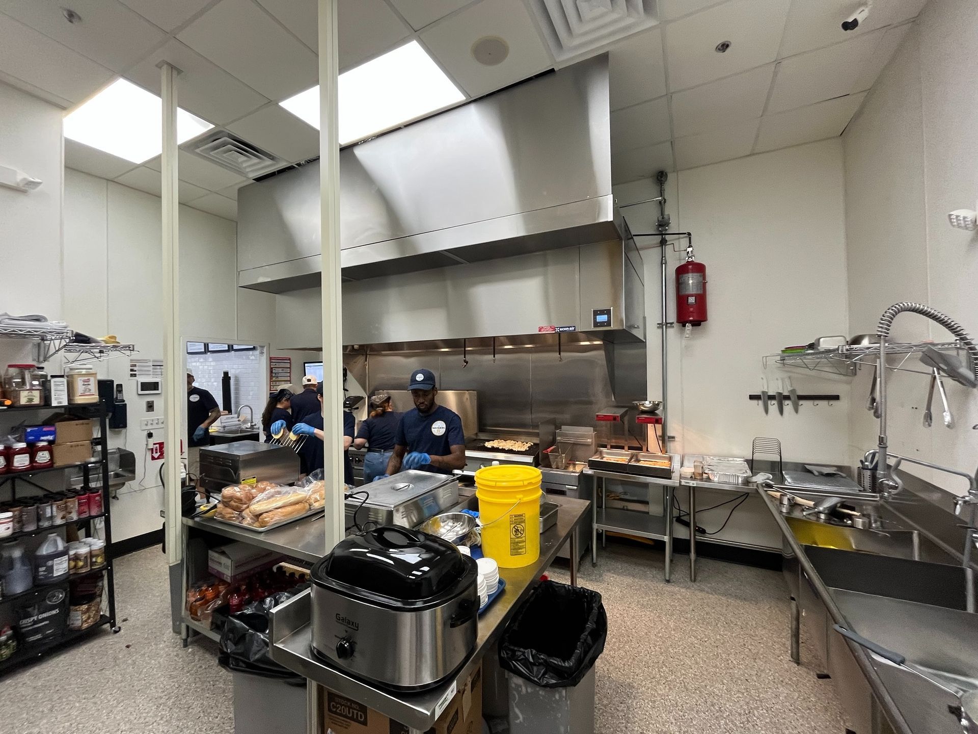 Commercial kitchen with cooking area, stainless steel appliances, and two workers preparing food.