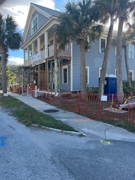 Two-story house under construction beside a sidewalk, with palm trees and orange safety fencing.