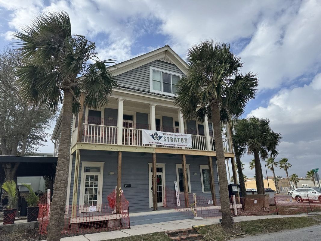 Two-story coastal house with a front porch, palm trees, and a banner, under a partly cloudy sky.