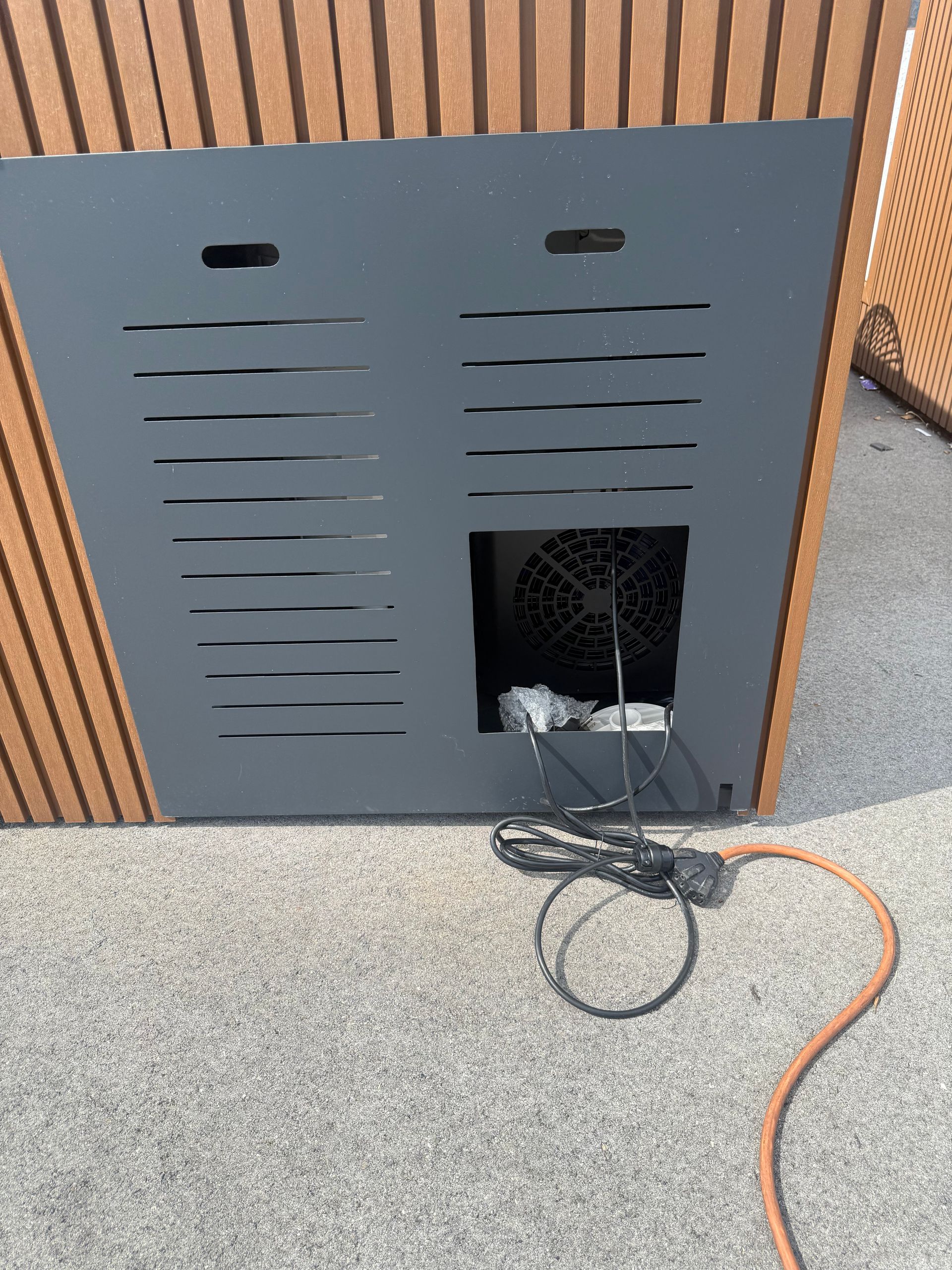 Gray vented panel on a wooden wall with an open lower access hole and an orange power cord on the floor.