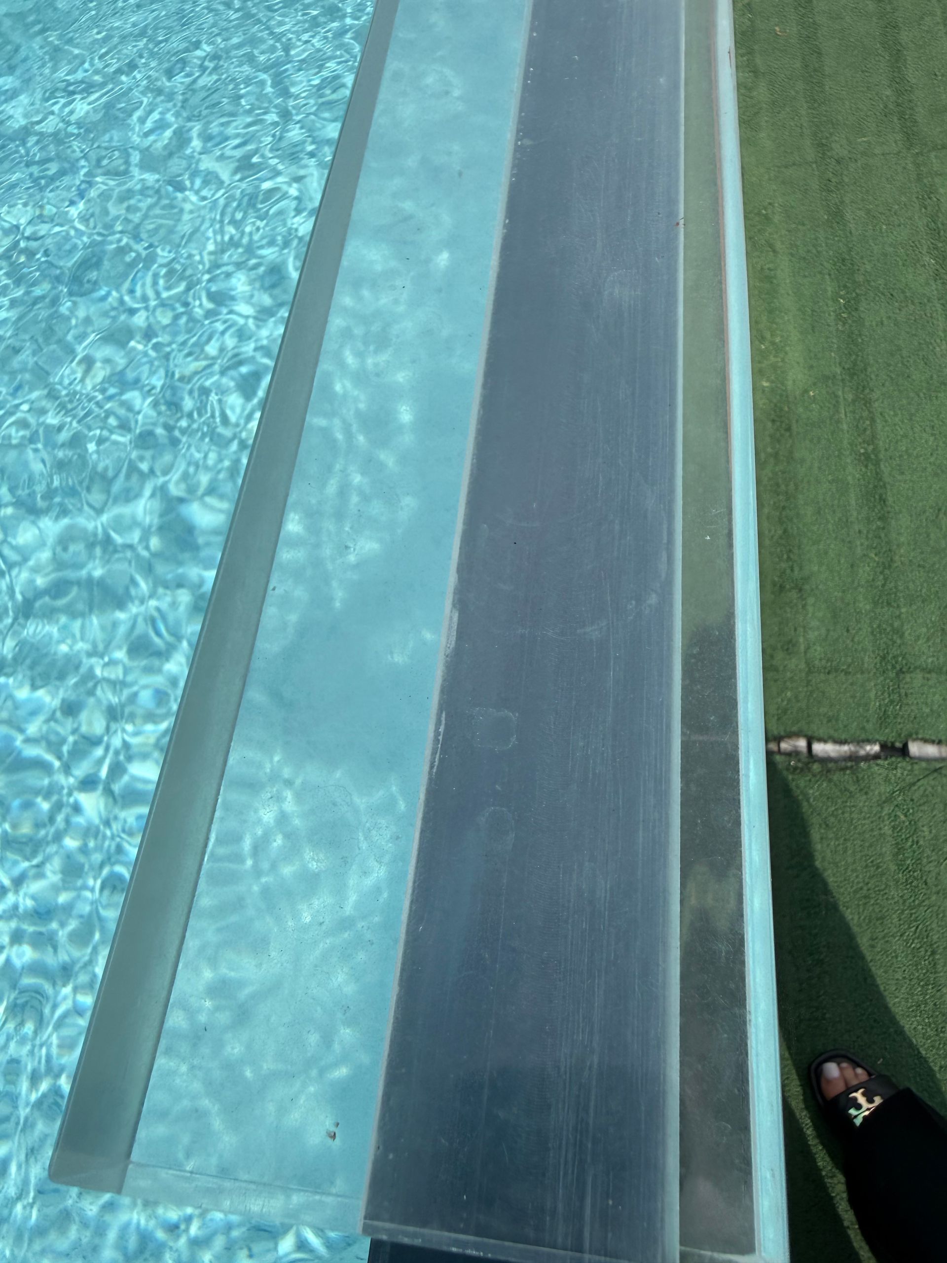 Pool ramp beside clear blue water and green turf, with a person’s foot at the lower right.