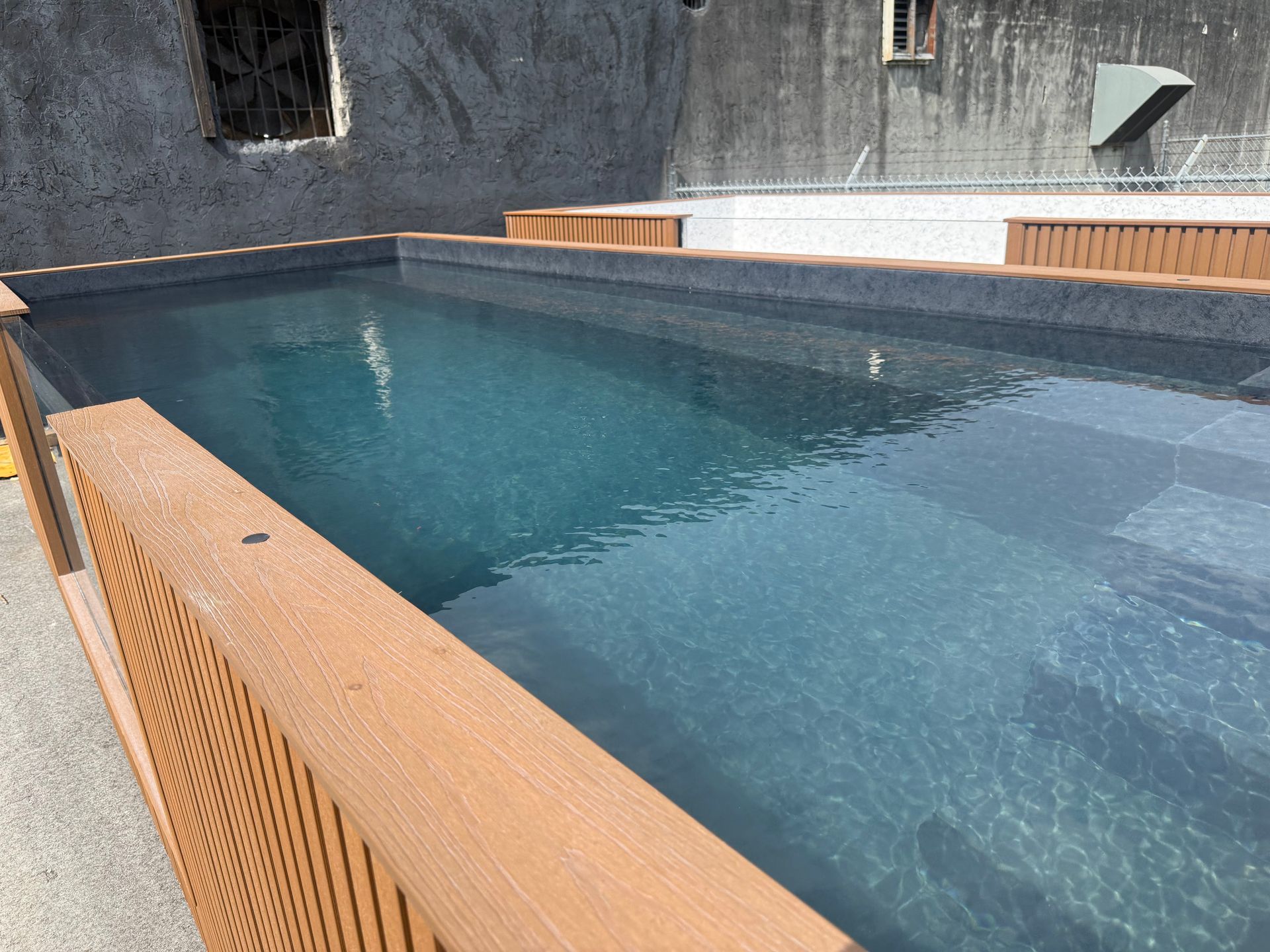 Blue swimming pool with wooden deck and stone wall in an outdoor courtyard