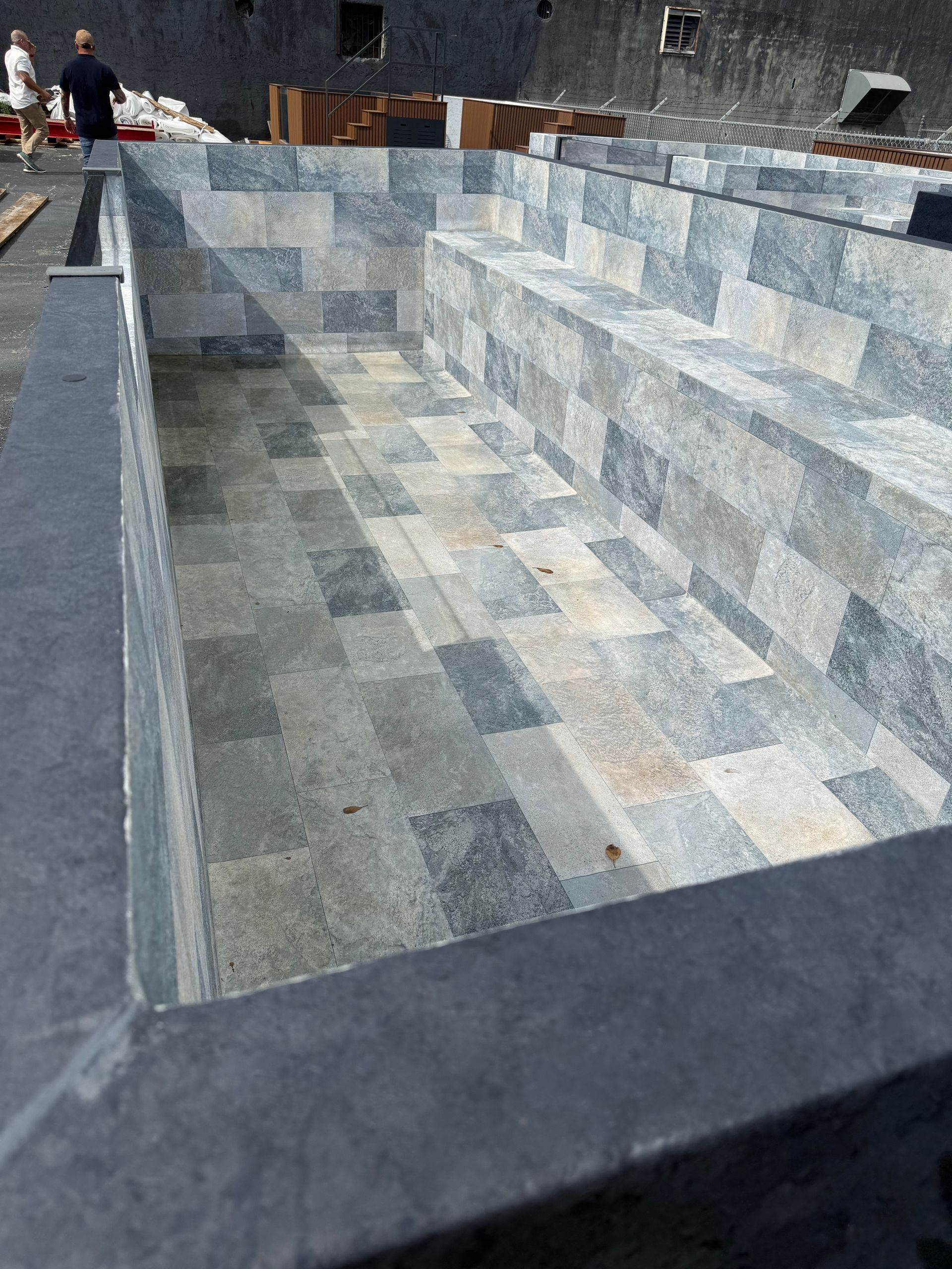 Large empty stone-lined pool or fountain with gray-blue tiled interior and a few people in the background