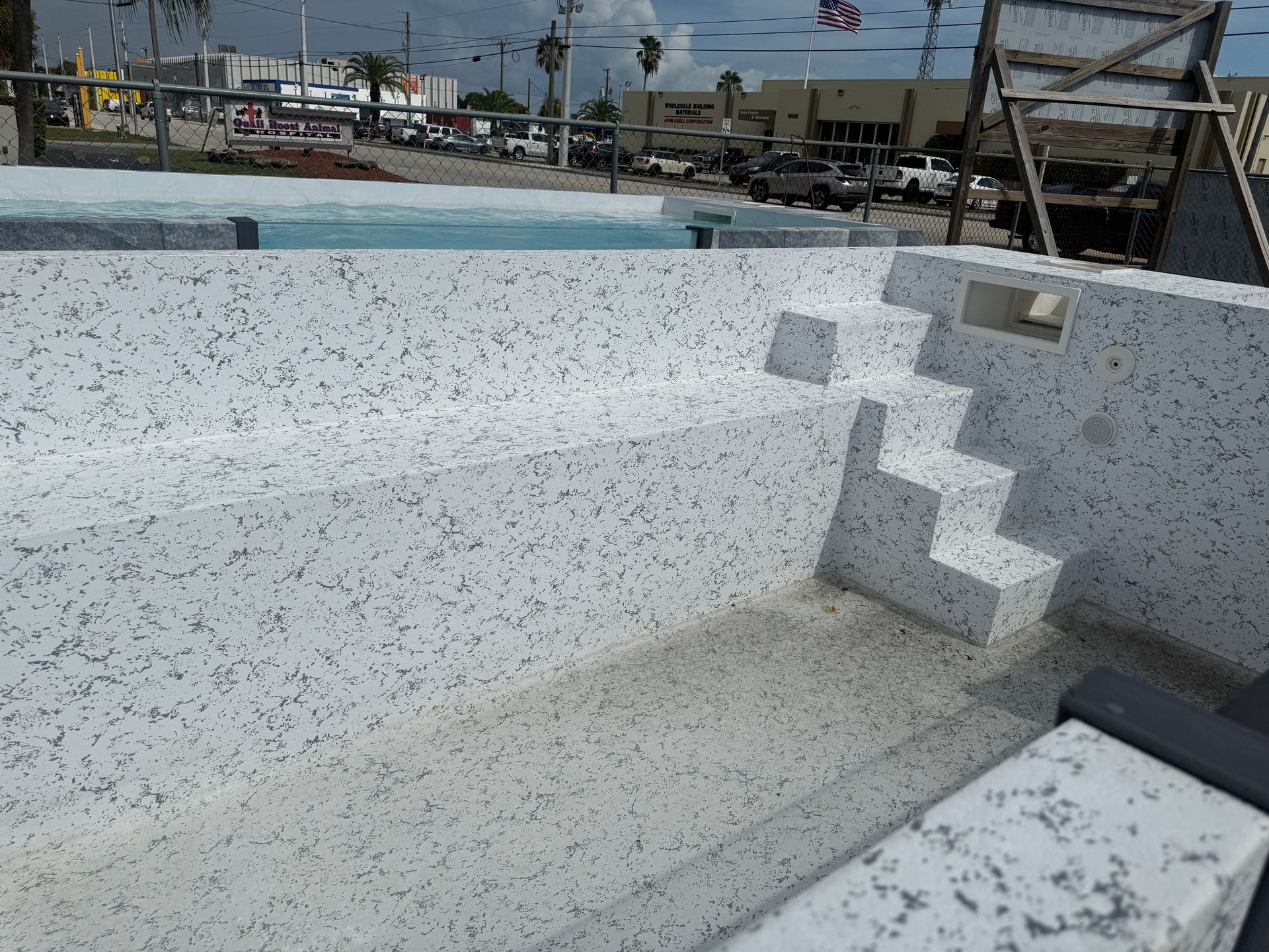 Empty speckled white swimming pool with built-in steps and deck chairs in the background