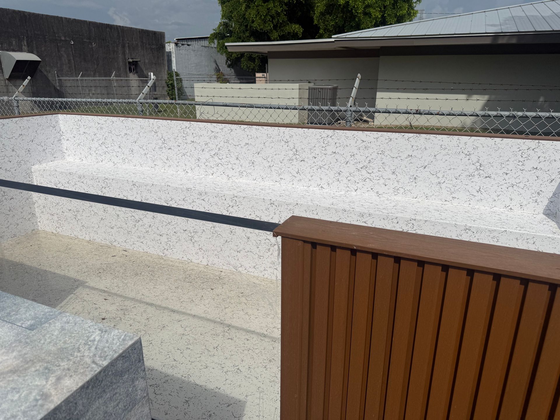 Outdoor pool with a white surface, gray deck, and brown slatted railing beside modern buildings.