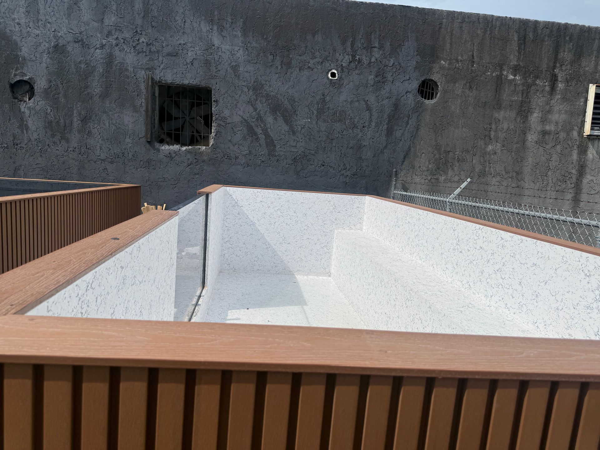 Empty white pool beside a concrete wall, framed by brown wooden decking