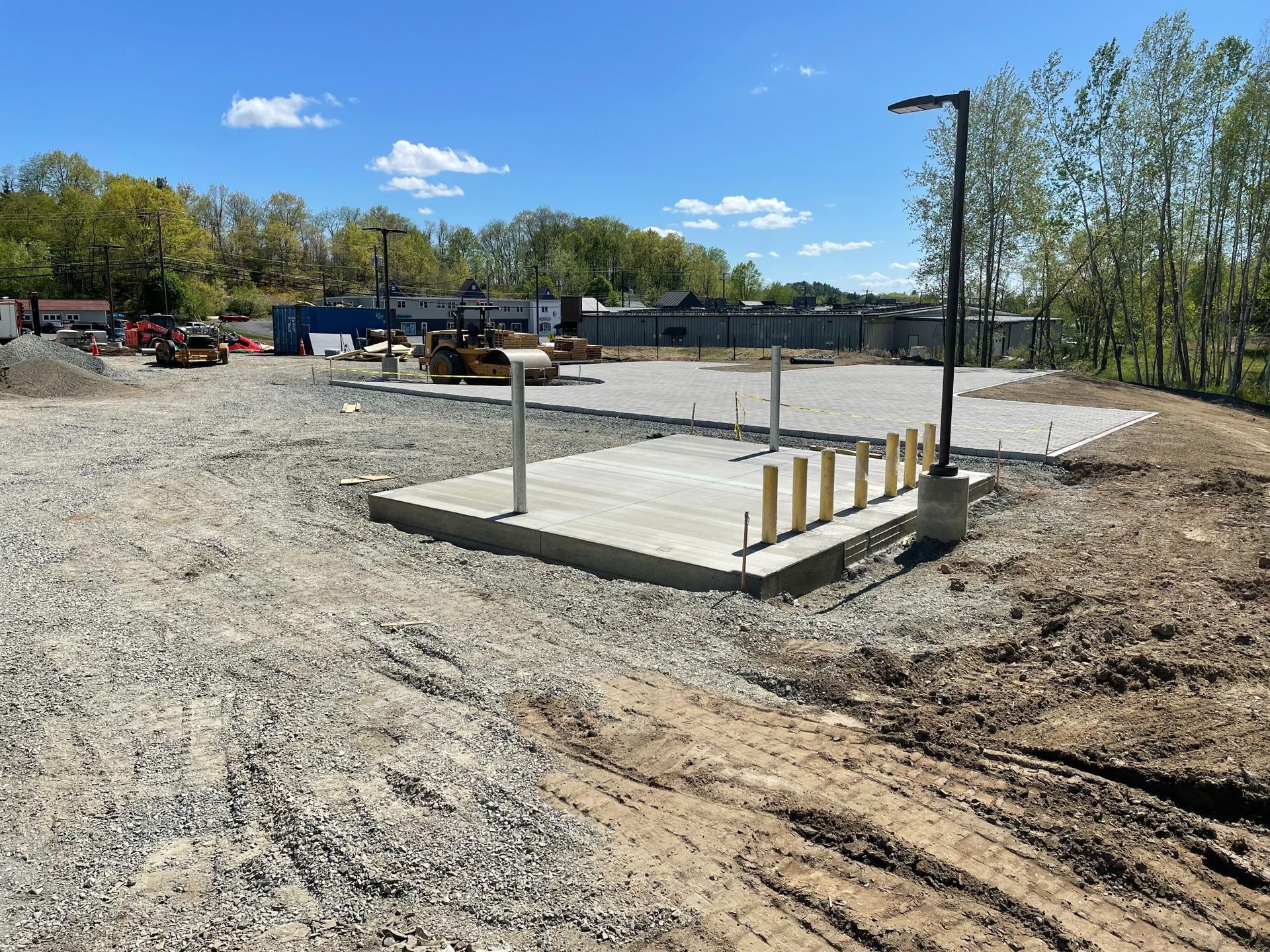 Construction site with concrete pads and gravel. Blue sky, trees, and heavy machinery in the background.
