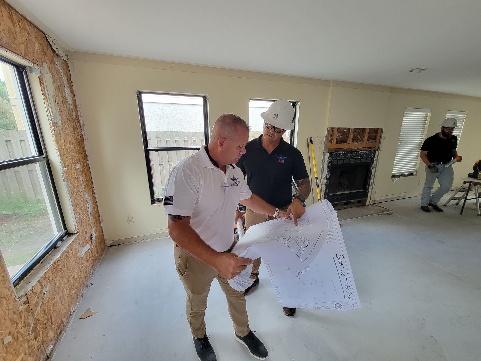 Two men in a room under construction examining blueprints. One wears a hard hat, the other a polo shirt.