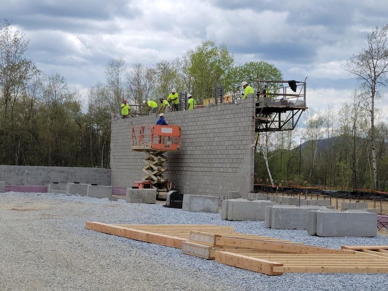 Construction workers build a concrete block wall with a lift platform, outdoors on a gravel lot under a cloudy sky.