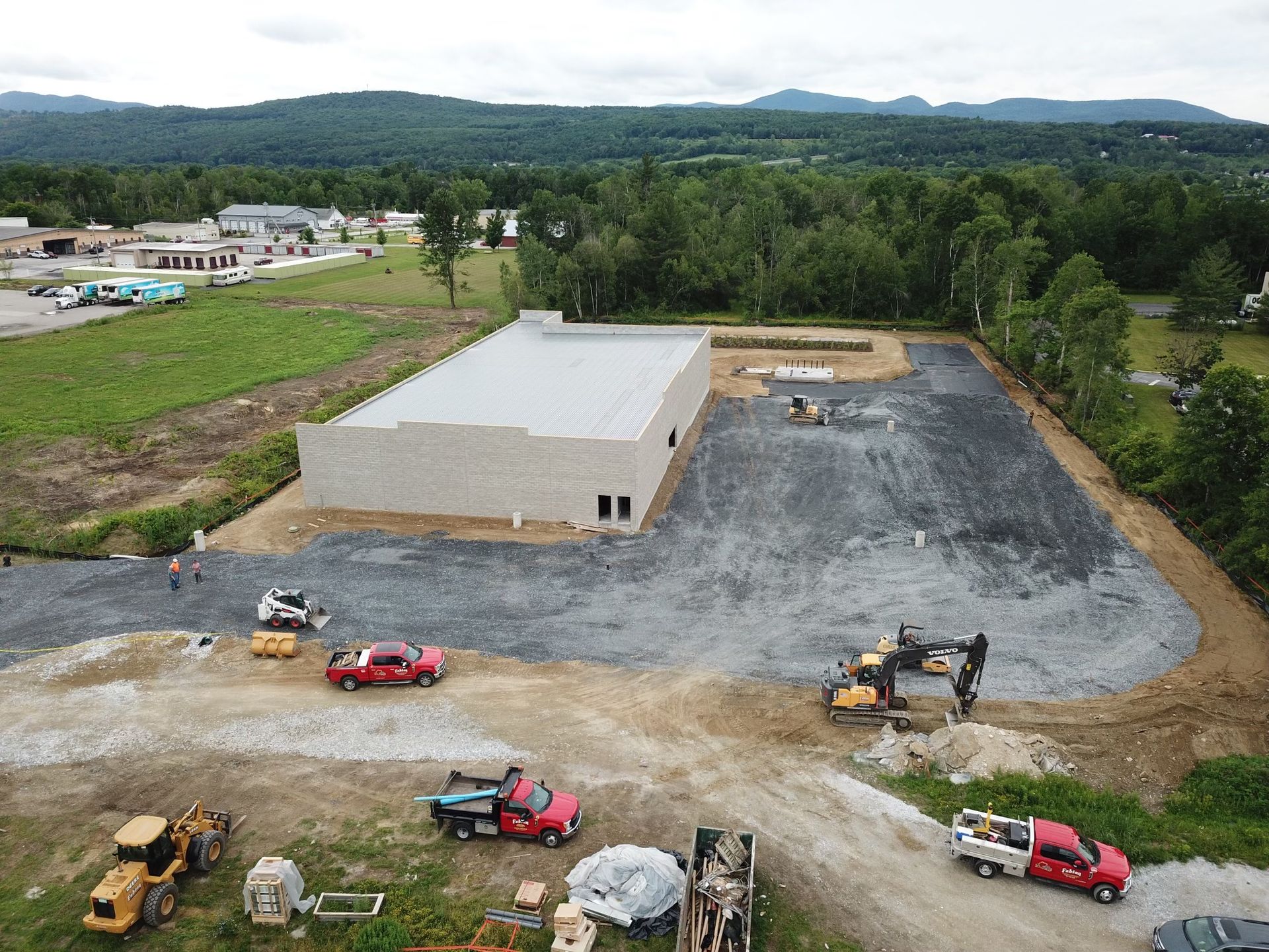 Construction site with a building, trucks, and heavy machinery, set against a backdrop of trees and mountains.