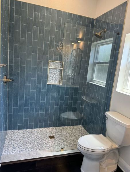 Blue-tiled shower with pebble floor, built-in shelf, and small window next to a white toilet in a bathroom.
