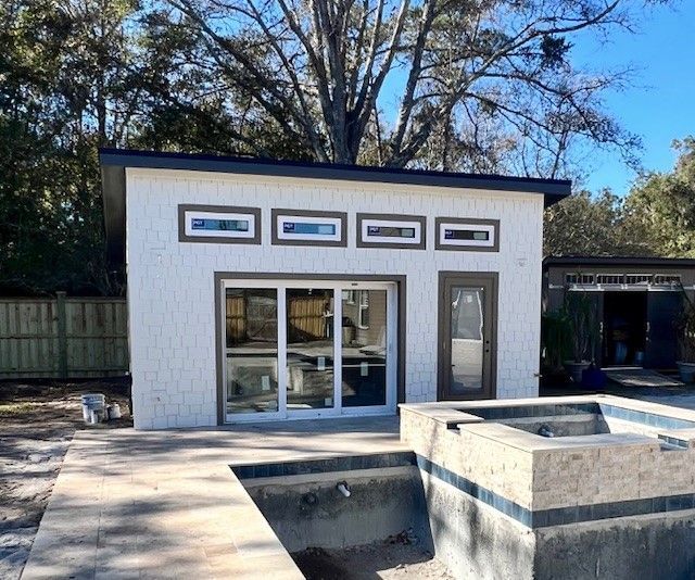 Modern white pool house with sliding glass doors, small rectangular windows, and a dark roof.